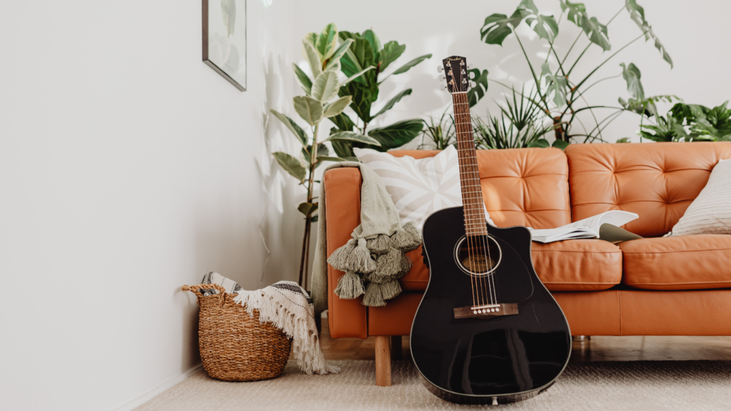 A room with a peach colored sofa and a black acoustic guitar standing up against the sofa.